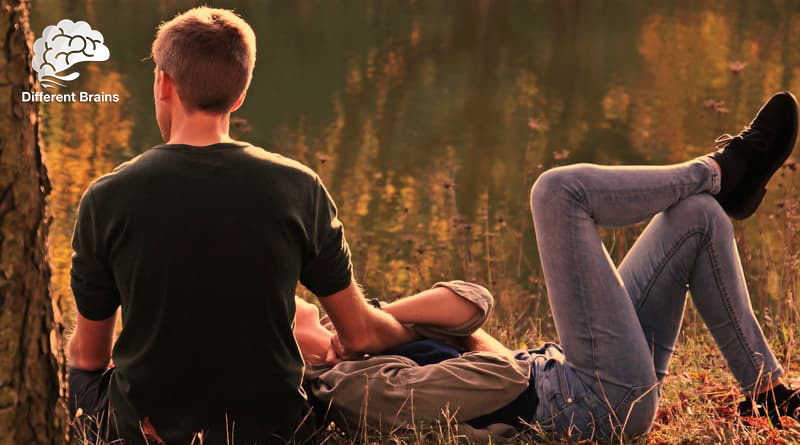 A woman lays on the ground in a field as a man sits up with his hand holding hers.