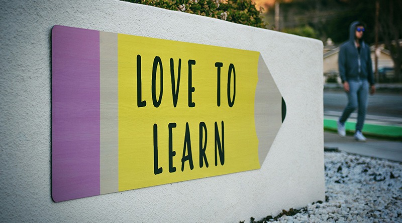 A Pencil-shaped Sign On A Short Outdoor Wall Reads "LOVE TO LEARN" While A Person Walks By In The Background