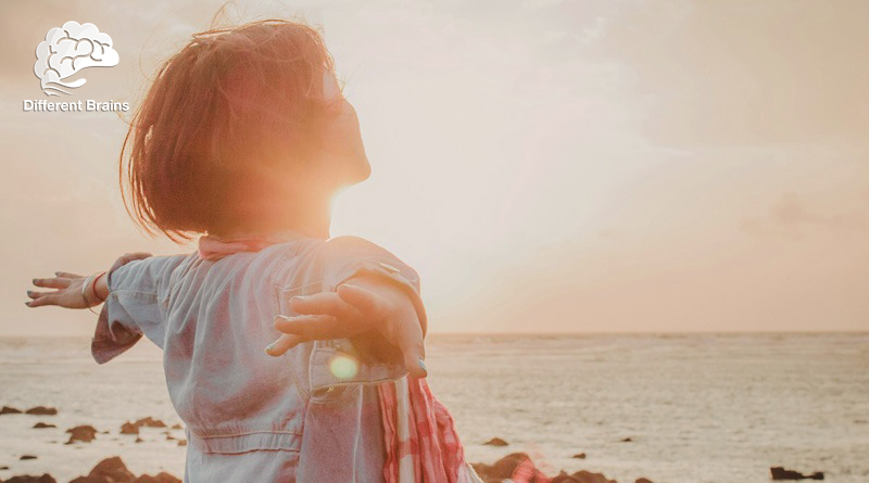 A woman stands on the beach looking in an orange sky with arms spread.