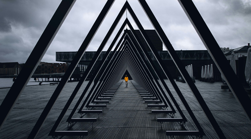 A person walks through a long triangular structure under an overcast sky.