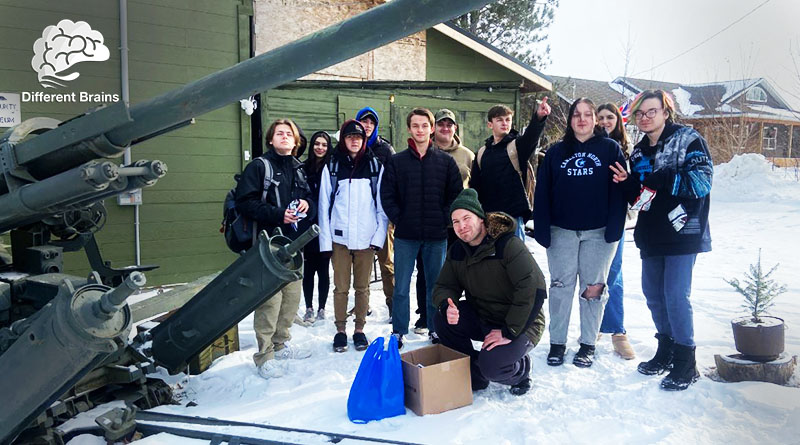 Aaron Bouma squats and gives a thumbs up with a group of students in front of the green exterior of the museum. Part of a tank is seen in the foreground.