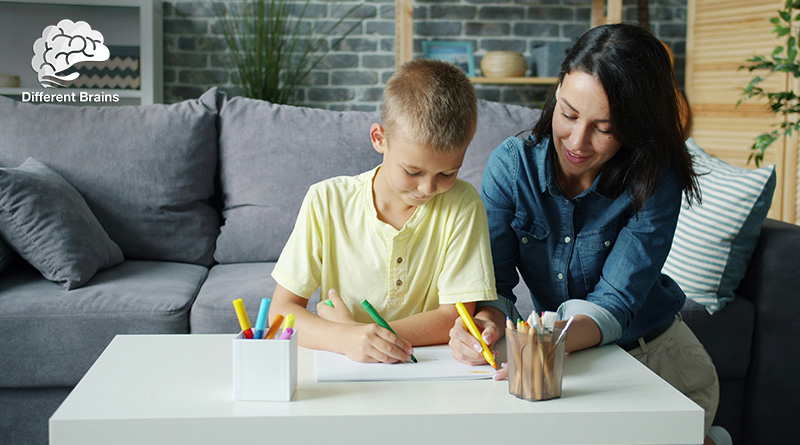 A woman leans next to her son and watches him writing.